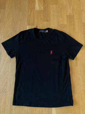 Svart t-shirt från Polo Ralph Lauren - Klassisk svart t-shirt från Polo Ralph Lauren med röd broderad logga på bröstet. T-shirten har rund halsringning och korta ärmar. Tillverkad i mjuk bomull för en skön och avslappnad stil. Skicka gärna vid funderingar. 