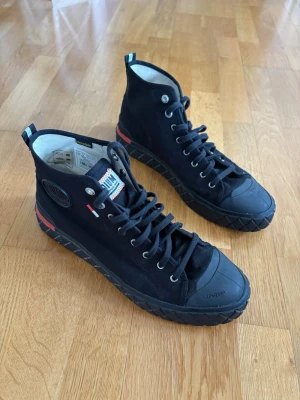 Svarta Palladium sneakers - Säljer ett par svarta sneakers från Palladium med hög canvasöverdel, robust gummisula och tåskydd. Skorna har svarta snören, metallöljetter och detaljer i rött, vitt och blått på plös och häl. Klassisk logga på sidan och streetinspirerad design.