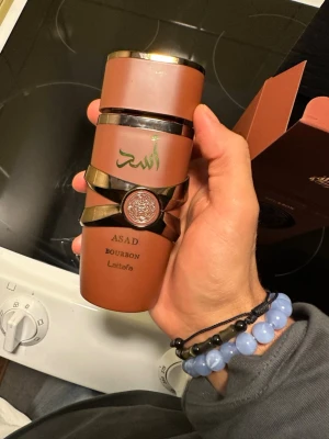 Lattafa Asad Bourbon parfym - Lattafa Asad Bourbon parfym i en brun flaska med gulddetaljer och arabiska inskriptioner. Flaskan har en unik design med ett dekorativt band och emblem i mitten. Kommer i matchande brun förpackning med guldfärgade texter. Flaska är helt nytt och oanvänd