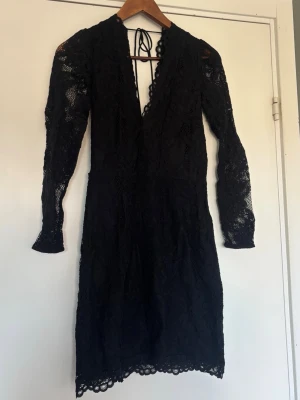 Svart spetsklänning med v-ringning - Svart klänning i spets med djup v-ringning och långa ärmar. Klänningen har knytning i nacken och vackra spetsdetaljer över hela plagget. 