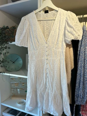 Vit kort klänning med broderade blommor - Superfin vit kort klänning med broderade blommor, puffärmar och v-ringning. Klänningen har knappar framtill och volang nertill. Perfekt för sommarens alla tillfällen och är gjord i luftig bomull.