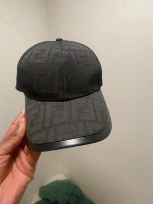 Svart keps från Fendi med FF-mönster - Snygg svart keps från Fendi med det klassiska FF-logomönstret över hela kepsen. Skärmen har en blank finish som ger en extra touch. Perfekt accessoar för att lyfta din outfit och visa stilkänsla.