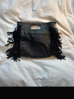 Svart handväska med fransar maje liknande - Snygg svart handväska i konstläder med fransar på sidorna och rektangulärt handtag. Väskan har även en avtagbar axelrem för olika stilar. Perfekt accessoar för dig som gillar boho-vibbar och vill sticka ut lite extra. Maje liknande väska