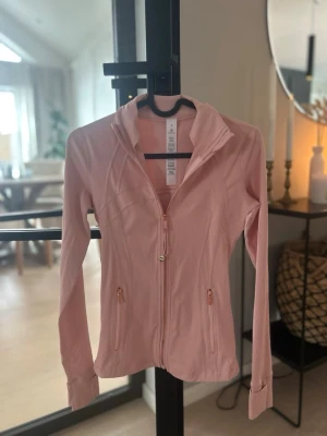 Ljusrosa träningsjacka med dragkedja - Snygg ljusrosa träningsjacka från Lululemon med hel dragkedja framtill och två fickor med dragkedja. Figurnära passform, hög krage och tumhål vid ärmsluten. Perfekt för gymmet eller löprundan.