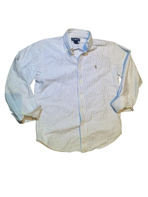 Randig skjorta från Ralph Lauren - Klassisk skjorta från Ralph Lauren med tunna blå och vita ränder. Skjortan har lång ärm, knappar framtill och en liten broderad logga på bröstet. Tillverkad i mjuk bomull och har en tidlös, normal passform.