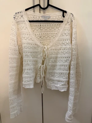 Vit virkad kofta från Aigou - Söt vit virkad kofta från Aigou med knytning framtill. Koftan har lång ärm, v-ringning och ett luftigt, spetsliknande mönster. Perfekt att slänga över en topp för en chill och trendig look.