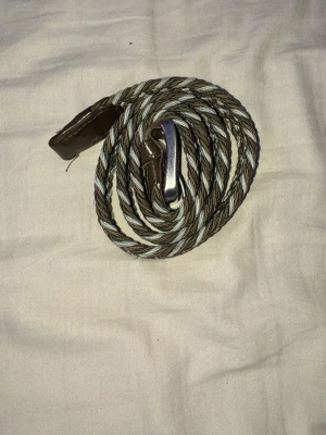 Brunt grått och vitt bälte  - Snyggt flätat bälte i bruna och grå toner med silverfärgat spänne. Bältet har detaljer i brunt skinn vid ändarna och ett mönster som ger en cool och modern look. Perfekt accessoar för att lyfta din outfit.