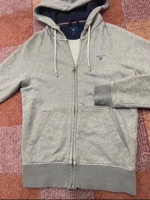 Grå zip-hoodie från Gant - Snygg grå hoodie från Gant med dragkedja, huva med dragsko och två stora fickor framtill. Mjuk insida och mörkblått foder i huvan. Broderad Gant-logga på bröstet. Perfekt för en chill och avslappnad stil.