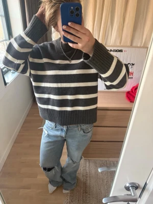 Randig stickad tröja i grått och vitt - Säljer en stickad tröja med breda horisontella ränder i mörkgrått och vitt. Tröjan har en avslappnad passform, ribbade muddar och nederkant samt rund halsringning. Perfekt för en chill och stilren look. Den är liten i storleken.