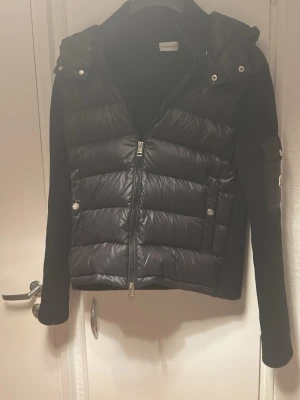 Svart pufferjacka från Moncler - Svart pufferjacka från Moncler med quiltad front, stickade ärmar och dragkedja. Jackan har en ficka med Moncler-logga på ena ärmen och snygga silverdetaljer. Perfekt för dig som vill ha en stilren och sportig look.