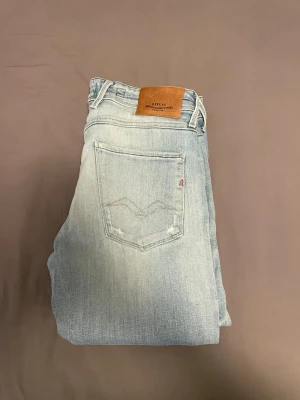 Ljusblå slitna jeans från Replay - Säljer ett par ljusblå jeans från Replay med slitningar och distressed detaljer. Klassisk femficksmodell med läderpatch bak i midjan och smal passform. Jeansen är tillverkade i mjuk denim och har en modern, avslappnad look.