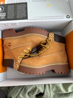Timberland klassiska kängor i nubuck - Säljer ett par ikoniska Timberland kängor i ljusbrun nubuck med grov sula och gulbruna snören. Kängorna har mörkbrunt vadderat skaft och tydliga sömmar. Perfekta för dig som gillar streetstyle och robust look.