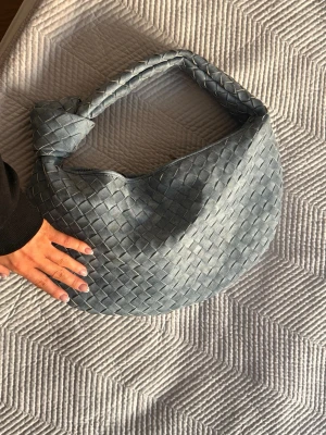 Blå flätad axelväska med knut - Snygg blå axelväska med flätad design och en unik knutdetalj på handtaget. Väskan har en mjuk, rundad form och stängs med dragkedja. Perfekt för dig som gillar trendiga accessoarer med lite extra edge.