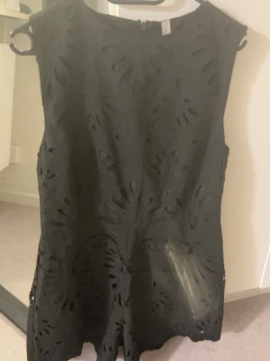 Svart broderad kortklänning - Snygg svart kortklänning med broderade detaljer och utskurna mönster. Klänningen är ärmlös och har rund halsringning. Materialet ser ut att vara bomull eller polyester och modellen är figurnära upptill med lätt utställd kjol.