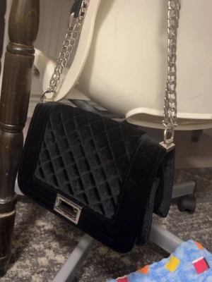 Svart quiltad axelväska med kedja - Snygg svart axelväska i sammet med quiltat mönster och silverfärgad kedjerem. Väskan har en rektangulär form och ett stilrent spänne framtill i metall. Perfekt för dig som vill ha en trendig och elegant accessoar till din outfit.