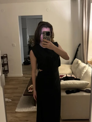 Svart långklänning med slits - Cool svart långklänning med ärmlös design och snygga cut-outs vid midjan. Klänningen har en slits framtill som ger extra edge och är perfekt för dig som gillar att sticka ut. Materialet känns mjukt och följsamt mot kroppen.