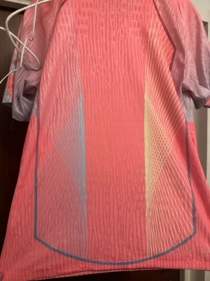 Rosa sporttröja med grafiskt mönster - Säljer en rosa sporttröja med korta ärmar och grafiskt mönster i blått, vitt och gult. Tröjan har en modern design med tunna linjer och gradienteffekt. Tillverkad i ett lätt och ventilerande funktionsmaterial som passar perfekt till träning eller löpning.