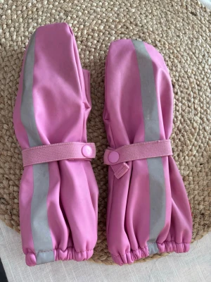 Rosa galon vantar  med reflexdetalj - Säljer ett par rosa vantar med breda reflexränder framtill och praktisk resår med knappstängning runt handleden. Perfekta för kalla och mörka dagar när du vill synas lite extra. Vantarna är långa och täcker en bit upp på armen. Storlek 3, 3-4 år 