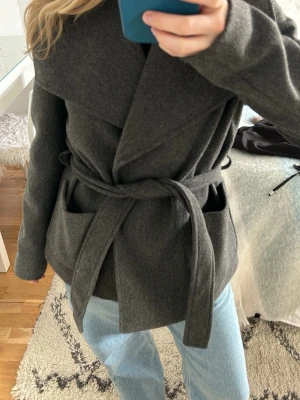 Mörkgrå kappa med knytskärp - Mörkgrå kappa med snöre som man kan ta av. Stora fickor med fina detaljer som passar perfekt till våren