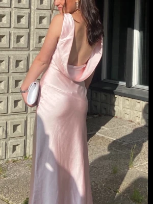 Ljusrosa långklänning med öppen rygg - Elegant ljusrosa långklänning i satin med v-ringad front och breda axelband. Klänningen har en snygg öppen rygg och ett mjukt fallande material som ger en lyxig känsla. Perfekt för speciella tillfällen. Finns inte att köpa längre på hemsidan! Använd endast en gång 