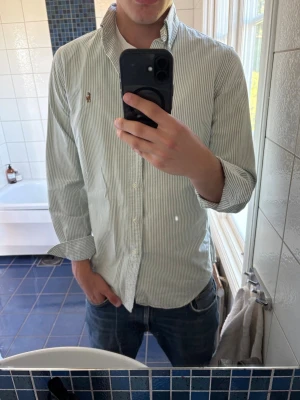 Randig skjorta från Polo Ralph Lauren - Klassisk skjorta från Polo Ralph Lauren med tunna vita och ljusgröna ränder. Skjortan har button-down krage, långa ärmar och broderad logga på bröstet. Tillverkad i bomull och har en normal passform. Perfekt till jeans eller chinos. Liten fläck vid bröstet men inget som syns tydligt vid användan.