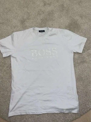 Vit Hugo Boss t-shirt med logga - Säljer en vit t-shirt från Hugo Boss med stor, vit logga på bröstet. Klassisk rund halsringning och korta ärmar. Tillverkad i mjuk bomull för en skön känsla. Perfekt till jeans eller shorts för en clean look.