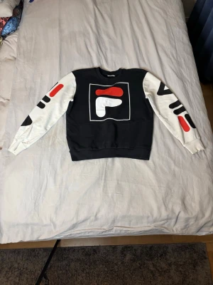 Sweatshirt från Fila - Cool sweatshirt i svart och vitt med stora grafiska tryck i rött, vitt och svart på både ärmar och bröstet. Tröjan har rund hals och ribbade muddar. Perfekt för dig som gillar streetwear och vill sticka ut med en unik design.