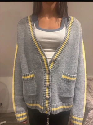 Grå kofta med gula detaljer - Mysig grå kofta med knappar framtill och två fickor. Koftan har gula och vita ränder längs kanterna, ärmsluten och nederkanten. Oversized passform och mjukt stickat material som ger en avslappnad look.