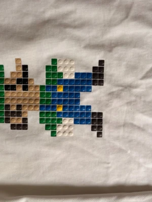 Vit t-shirt med pixelmönster - Vit t-shirt med coolt pixelinspirerat tryck i blått, grönt, svart, beige och gult på bröstet. Rund halsringning med texten 'HONE BABY' på insidan. Perfekt för dig som gillar retrostil och unika detaljer.