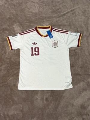 Vit Spanien fotbollströja Adidas - Säljer en vit Spanien fotbollströja från Adidas med vinröda och gula detaljer. Tröjan har nummer 19 och namnet Lamine Yamal på ryggen, samt landslagets emblem på bröstet. Klassisk sportdesign med korta ärmar och rund hals. Perfekt för match eller träning.