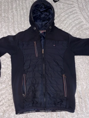 Marinblå vindjacka Tommy Hilfiger - Snygg marinblå vindjacka från Tommy Hilfiger med huva, dragkedja framtill och flera fickor med dragkedja. Jackan har quiltade partier och diskret logga på bröstet. Perfekt för blåsiga dagar och har sportig look med detaljer i rött, vitt och blått. Dock är en ficka sönder.