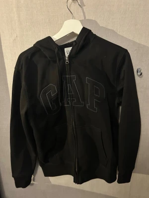 Svart zip hoodie från GAP - Säljer en svart hoodie från GAP med dragkedja framtill och stor logga broderad på bröstet. Hoodien har huva med dragsko och två fickor fram. Tillverkad i mjukt sweatshirtmaterial, perfekt för chill dagar.