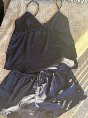 Mörkblå satinset med linne och shorts - Tvådelat set i mörkblå satin med ett linne med smala axelband och v-ringning samt matchande shorts med resår i midjan och volangkant. Materialet är glansigt och mjukt, perfekt för en avslappnad känsla.