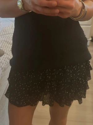 Svart volangkjol med vita prickar - Snygg svart kortkjol med vita prickar och flera volanger. Shorts under kjolen, dragkedja på sidan av kjolen. I mycket bra skick!