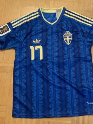 Blå Sverige fotbollströja nr 17 - Säljer en blå fotbollströja med gula detaljer från Sveriges landslag. Tröjan har nummer 17 och namnet Gyökeres på ryggen, samt Sveriges landslagsemblem och Adidas-logga på bröstet. Klassisk retrodesign med mönster och gula ränder på axlarna.
