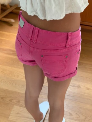 Rosa shorts med upprullad kant - Snygga rosa shorts med upprullad kant och klassisk femficksdesign. Shortsen har bälteshällor och silverfärgade nitar vid fickorna. Perfekta för varma dagar och ger en fräsch look.