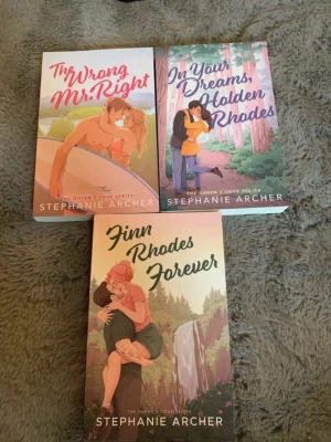 The Queen's Cove-serien av Stephanie Archer - Säljer tre romantiska pocketböcker av Stephanie Archer: 'The Wrong Mr. Right', 'In Your Dreams, Holden Rhodes' och 'Finn Rhodes Forever'. Omslagen har färgstarka illustrationer i rosa, lila och beige toner. Perfekt för dig som gillar feelgood och kärlekshistorier.