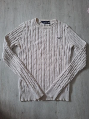 Vit kabelstickad tröja GANT - Kabelstickad tröja i vitt från GANT med rund halsringning och diskret logga broderad på bröstet. Långa ärmar och ribbade muddar vid ärmslut och nederkant. Klassisk och stilren design som passar till mycket.