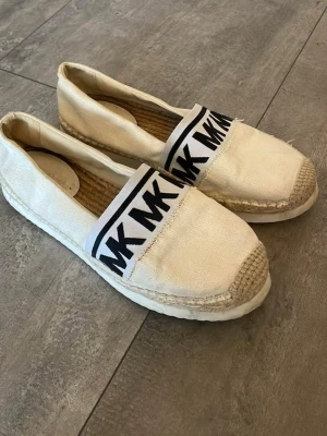 Vita espadrillos MK - Snygga vita ballerinaskor i espadrille-stil från Michael Kors. Skorna har en bred resår med svart MK-logga över foten och naturfärgad flätad sula. Ovandelen är i textil och sulan har en klassisk repdetalj. Perfekta för en avslappnad och trendig look.