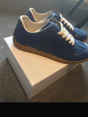 Blå sneakers Maison Margiela - Snygga blå sneakers från Maison Margiela med vita skosnören och beige gummisula. Skon är i mocka och textil, Och sulan har ett klassiskt mönster. Perfekta för dig som vill ha en clean och stilren look. Box följs med