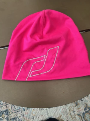 Neonrosa mössa med tryck - Snygg neonrosa mössa i stretchigt material med ett vitt grafiskt tryck på framsidan. Mössan har en enkel, slimmad passform och är perfekt för dig som vill sticka ut. Passar bra till både sport och streetwear. Har lite små fläckar någonstans som inte syns tydligt