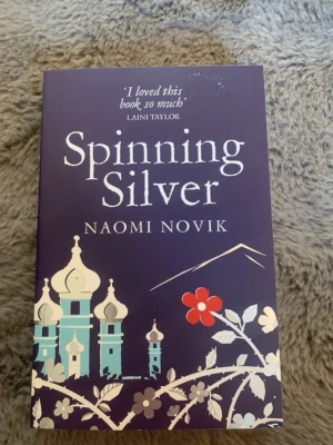 Spinning Silver av Naomi Novik - En engelskspråkig fantasybok med lila omslag och dekorativa illustrationer av slott och blommor. Boken är skriven av Naomi Novik och har citat från kända författare på baksidan. Perfekt för dig som gillar magi, äventyr och starka kvinnliga huvudpersoner.
