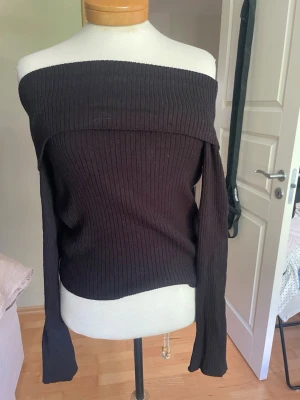 Svart ribbad offshoulder topp - Snygg svart ribbad topp med offshoulder och långa ärmar. Toppen har en kroppsnära passform och är tillverkad i ett stretchigt stickat material som ger en modern siluett. Perfekt för dig som gillar stilrena och trendiga plagg.