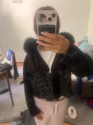 Svart flis med äkta päls  luva från alessa  - Mysig svart flis/hoodie med dragkedja och stor fluffig luva. Jackan är kort i modellen och har en mjuk, lurvig yta som ger en trendig look. Perfekt för dig som vill ha en varm och stylis.