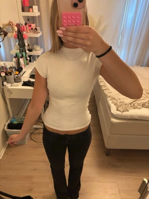 Vit ribbad polotopp - Säljer en vit ribbad polotopp med korta ärmar och tight passform. Toppen har en hög krage och är tillverkad i ett mjukt bomullsmaterial som sitter snyggt på kroppen. Perfekt att styla med jeans eller kjol.