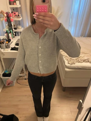 Ljusgrå cropped kofta med knappar - Säljer en ljusgrå cropped kofta med rund hals och vita knappar framtill. Koftan har långa ärmar och ribbade muddar vid ärmslut och nederkant. Perfekt att slänga över en topp för en chill look.