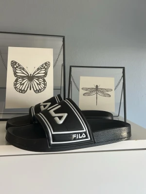 Svarta Fila badtofflor med vit logga - Säljer ett par svarta Fila badtofflor med bred rem och vit logga framtill. Tofflorna har en klassisk slides-design med två vita ränder längs remmen och är tillverkade i syntetmaterial. Perfekta för chill dagar eller till stranden.