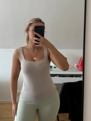 Beige  tanktop med fyrkantig ringning - Säljer en beige rtanktop med breda axelband och fyrkantig ringning. Toppen är figurnära och har en enkel, stilren design som passar perfekt till jeans eller kjol. Materialet ser mjukt och stretchigt ut för en bekväm passform.