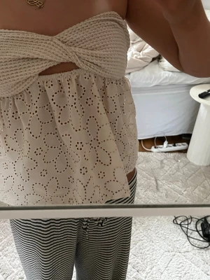 Beige broderad bandeau topp - Supersöt bandeau topp i beige med virkad twistad överdel och broderade blommor på den luftiga nederdelen. Perfekt för varma dagar och ger en romantisk vibe. Toppen är axelbandslös och har en liten cut-out framtill.