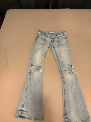 Ljusblå utsvängda slitna jeans - Säljer ett par ljusblå jeans med utsvängda ben och slitningar på både lår och knän. Jeansen har stora framfickor och klassisk femficksdesign. Perfekta för dig som gillar en avslappnad och trendig stil.
